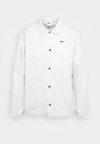 Vit knappar up Nike-jacka med krage, två framfickor och en liten svart Nike-swoosh-logotyp på bröstet.