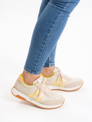 Person wearing beige and yellow sneakers with peach accents and blue jeans, standing on a white background.