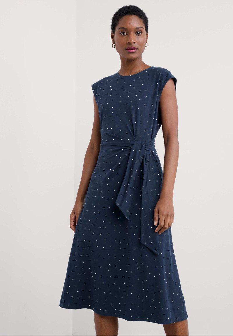 Robe bleu marine à manches courtes, détail de nœud à la taille, et pois blancs sur l'ensemble. Design fluide, de longueur mi-longue, texture de tissu douce.