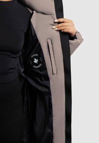 Abrigo acolchado con una capa exterior negra, forro marrón claro y bolsillo interior. Presenta una marca sutil y una textura suave.