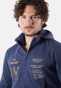 Giacca zip-up blu navy in tessuto morbido, con toppe ricamate, cappuccio e rifiniture a contrasto. Il testo include "AERONAUTICA MILITARE."