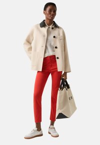 Cremefarbener Mantel mit schwarzem Kragen, rote Hose, weiße Turnschuhe und eine beige Tragetasche mit schwarzen Akzenten. Weiche Textur und strukturierte Form.