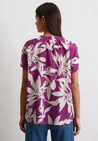 Lila Bluse mit floralem Muster und elfenbeinfarbenen Blättern, kurzen Ärmeln, gerafftem Ausschnitt und lockerer Silhouette, kombiniert mit Jeans.