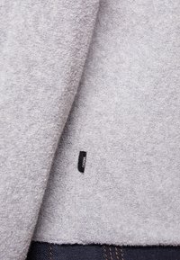 Grauer Fleece-Stoff mit weicher Textur, ausgestattet mit einem angehängten schwarzen Etikett. Der Saum ist gegen dunkelblauem Denim mit orangefarbenen Nähten sichtbar.