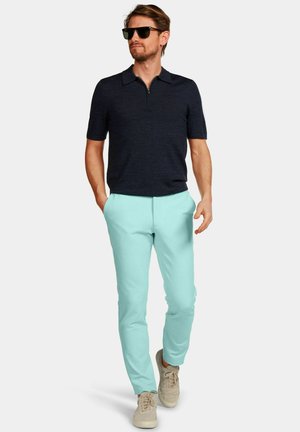 Man met zwarte korte mouwen polo, lichtblauwe broek, beige sneakers en zwarte zonnebril, lopend met handen in de zakken tegen een witte achtergrond.