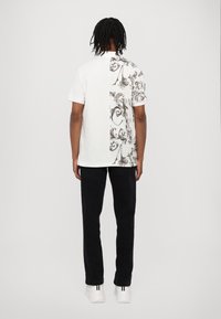 Balta T-kreklā ar rakstainu pelēku akcentu vienā pusē, kurā ir sarežģīti zīmējumi; savienota ar melnām biksēm un baltām kurpēm.