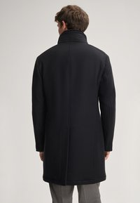 Czarny wełniany płaszcz z wysokim kołnierzem, bocznymi kieszeniami i eleganckim krojem. Gładka tekstura i dopasowany fason, kończący się tuż nad kolanami.