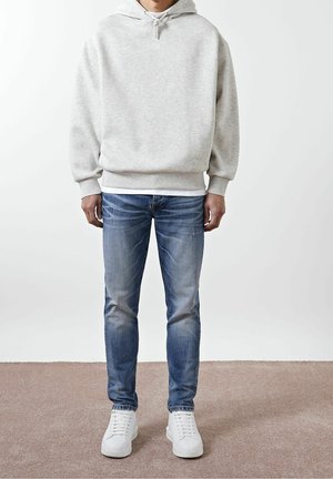 Osoba ubrana w jasnoszarą bluzę z kapturem, niebieskie wyblakłe dżinsy i białe trampki stoi na jednolitym tle.