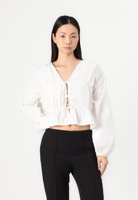 Blusa bianca cropped con scollatura a V, vita arricciata e maniche lunghe e voluminose. Presenta un dettaglio con lacci sul petto e una texture leggera.