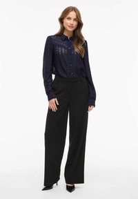 Marineblå blondebluse med knaplukning, der har et mønstret design, parret med høj talje sorte vide bukser og sorte højhælede sko.