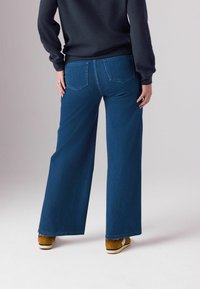 Jeans din denim albastru cu picioare largi și talie înaltă, cu două buzunare pe spate și detalii de cusătură minime. Purtați cu adidași maroon.