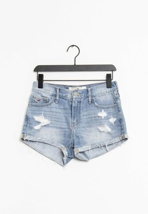 Short en denim bleu clair délavé avec des ourlets effilochés suspendu à un cintre en plastique noir sur fond blanc.