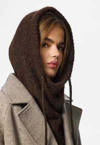 Brun stickad halsduk med långa knytband, dragen under en ljusbrun texturerad kappa, som ramar in ett ansikte med rakt blondt hår.