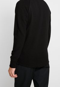 Camisa negra de manga larga con textura acanalada, cuello redondo estándar y detalles en los puños en contraste. Diseño simple sin gráficos ni adornos.