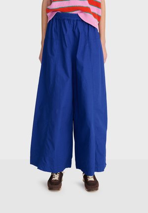 Persoon die felblauwe wijde broek, een roze-oranje gestreepte trui en bruine sneakers met witte veters draagt.