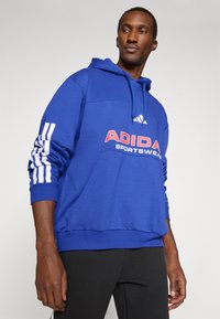 Modra Adidas kapuca z kenguru žepom, kapuco na vrvico in belimi navpičnimi črtami na rokavih. Sprednji logotip nosi napis "ADIDAS SPORTSWEAR."