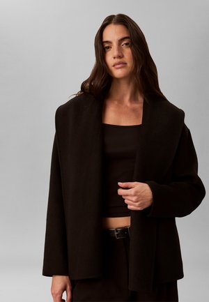 Kvinde med langt mørkt hår iført en løs sort jakke over en sort crop top, stående mod en lysegrå baggrund.