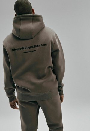 Brązowa bluza z kapturem i pasujące spodnie, wykonane z miękkiego materiału, z dużym czarnym logo z tyłu przedstawiającym "DiverseExtremeTeam.com."