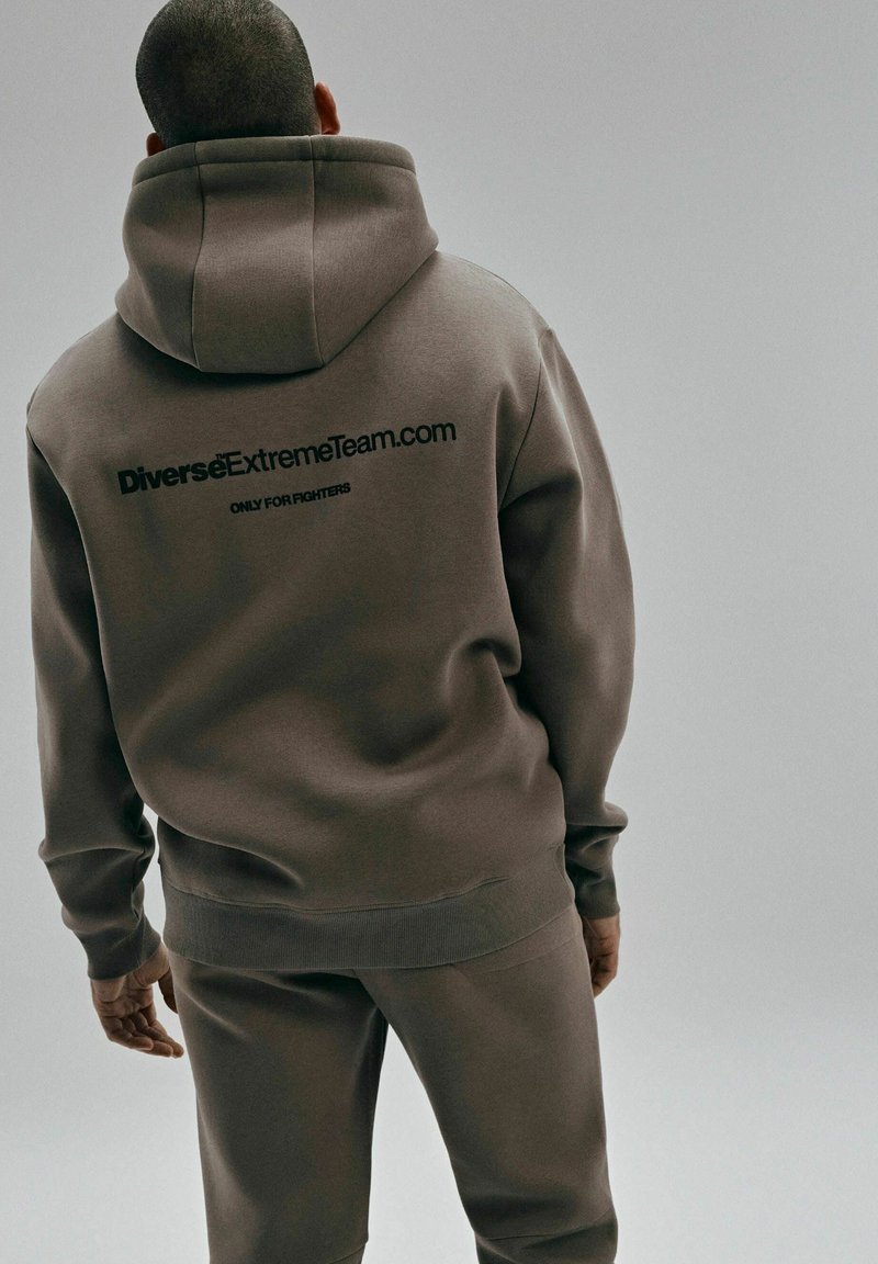 Brązowa bluza z kapturem i pasujące spodnie, wykonane z miękkiego materiału, z dużym czarnym logo z tyłu przedstawiającym "DiverseExtremeTeam.com."