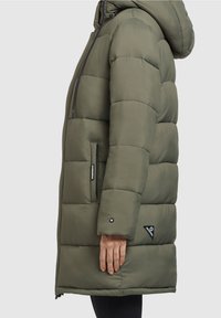 Giacca a piumino verde oliva con cappuccio, caratterizzata da una trama trapuntata, chiusura con cerniera e due tasche laterali con dettagli neri.
