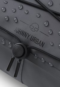 Borsa impermeabile grigia con gocce d'acqua sulla superficie, dotata di un toggle nero rotondo e cordoncino. L'etichetta riporta "JOHNNY URBAN" in nero.