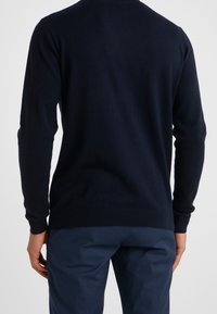 Marinblå stickad tröja med ribbade muddar och nederkant, med en enkel rund halsringning, visad från ryggen med en figursydd design.