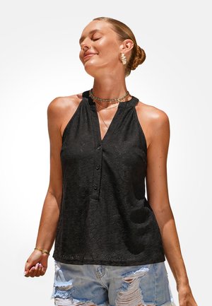 Vrouw met haar in een knot draagt een zwarte mouwloze top, gelaagde kettingen, oorbellen en gescheurde denim shorts, glimlachend met gesloten ogen.
