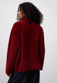 Giacca oversized bordeaux, morbida, con colletto alto e tessuto strutturato. Presenta una piccola etichetta nera sul retro.