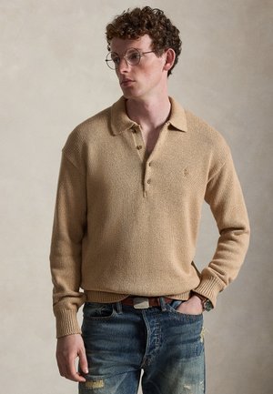 Jeune homme aux cheveux bouclés et lunettes rondes, portant un pull en tricot beige et un jean bleu déchiré, une main dans la poche, regardant sur le côté.