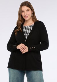Žena oblečená v černém kardiganu na knoflíky přes houndstooth top a modré džíny, s kruhovými náušnicemi, stojící na jednobarevném světlém pozadí.
