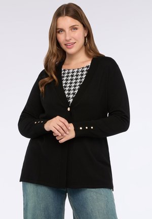 Vrouw in een zwarte knoopcardigan over een houndstooth-top en blauwe jeans, met hoepeloorbellen, staand tegen een effen lichte achtergrond.