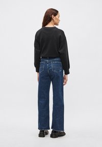 Čierna mikina spojená s modrými džínsami s vysokým pásom a širokými nohavicami. Džínsy majú zadné vrecká s kontrastným prešívaním; modelka má na sebe hrubé čierne topánky.