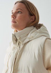 Hellbeige Steppweste mit Kapuze, geprägt von einem gesteppten Textil, Zugbänder und einem Reißverschluss vorne, getragen über einem gestrickten Pullover.