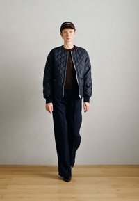 Quiltota melna bomber jakna virs brūna kardigāna, savienota ar tumši zilo svītraino biksēm un brūnu cepuri. Koka grīda, neitrāla fona.