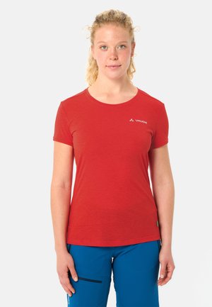 Vrouw met krullend blond haar, gekleed in een rood Vaude-shirt met korte mouwen en een blauwe broek, staand tegen een effen witte achtergrond.