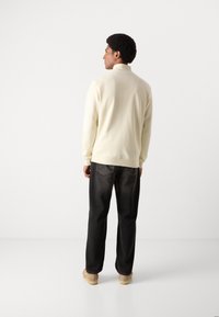 Lys gul pullover med høj krave, ribbede manchetter, struktureret stof; parret med mørke, afslappede jeans og beige fodtøj.