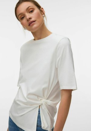 Femme portant un haut blanc à manches courtes avec un nœud sur le côté, associé à un jean bleu, posant devant un fond clair uni.