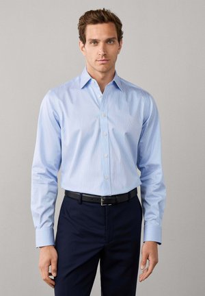 Man met kort bruin haar, die een lichtblauw overhemd draagt dat in een donkere broek met een zwarte riem is gestopt, staand tegen een effen achtergrond.