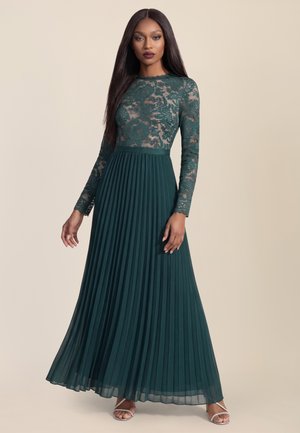 Donna con capelli lunghi e lisci che indossa un abito con top in pizzo verde scuro, gonna plissettata e tacchi argentati con cinturini, in piedi davanti a uno sfondo beige.