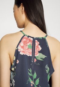 Blusa azul marinho com estampado floral, apresentando um detalhe em abertura nas costas, alças finas e um tecido suave e texturizado com padrões vibrantes em verde e rosa.