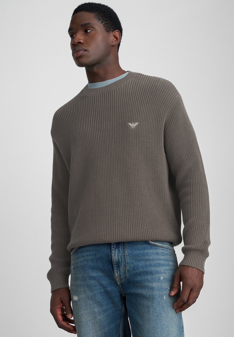 Rjav rebrast pulover s krošnjim ovratnikom in diskretnim logotipom na prsih, v kombinaciji s svetlo modrimi raztrganimi kavbojkami. Sproščen dizajn in tekstura.