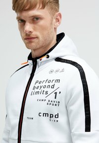Felpa bianca con zip e accenti neri e testo stampato. Presenta un cappuccio con coulisse, tasche laterali e elementi di design sportivi.