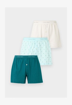 Trois boxers homme Calvin Klein en crème, bleu clair avec motif logo et couleur sarcelle foncée, disposés en chevauchement sur un fond blanc.