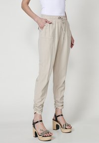 Beige drawstring pants dengan saku samping dan cuff elastis, dipadukan dengan sandal wedge bergaris hitam dan putih yang menampilkan detail tali anyaman.