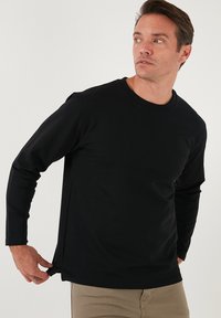 Maglietta nera a maniche lunghe in design aderente. Realizzata in tessuto liscio ed elasticizzato con scollatura rotonda e fessure laterali, indossata sopra pantaloni beige chiari.