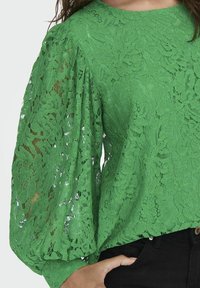 Blouse en dentelle verte avec manches bouffantes et motifs floraux, associée à un pantalon noir, main posée près de la poche.