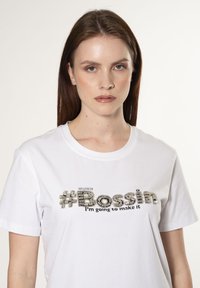Hvid bomulds t-shirt med et tekstureret grafisk design, hvor der står "#Bossin" i sølv og sort, samt teksten "Jeg vil klare det."