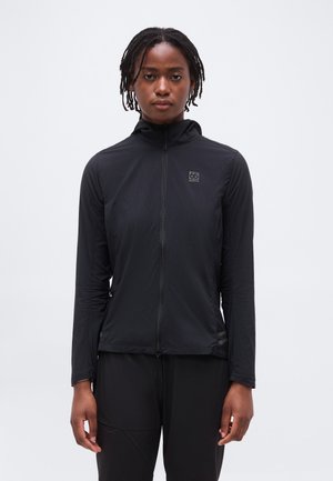 Personne avec de courtes dreadlocks portant une veste noire zippée avec le logo 66 North, debout devant un fond clair uni.