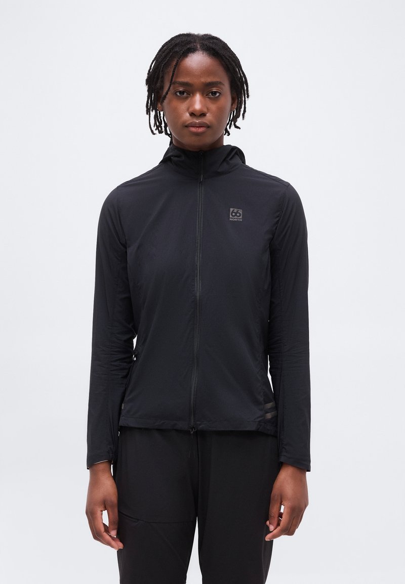 Personne avec de courtes dreadlocks portant une veste noire zippée avec le logo 66 North, debout devant un fond clair uni.