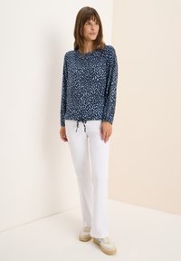 Blusa blu con un modello a macchie unico, maniche lunghe e un cordoncino all'orlo, abbinata a pantaloni bianchi e sneakers beige.
